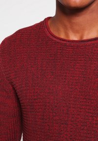 Punainen neulepaita, jossa on teksturoitu kuvio ja pyöreä kaula-aukko. Materiaali näyttää pehmeältä ja hieman joustavalta, ja siinä on tumma marle-efekti.