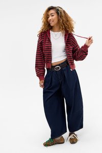 Νεαρή γυναίκα με σγουρά μαλλιά, που φοράει κόκκινο και μαύρο ριγέ φούτερ με κουκούλα, λευκό crop top, φαρδιά σκούρα τζιν και ασύμφωνες αθλητικές μπότες.