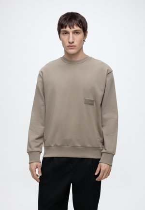 Uomo con capelli corti e scuri che indossa una felpa beige e pantaloni neri, in piedi davanti a uno sfondo chiaro uniforme.