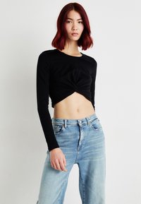 Crni dugih rukava crop top s dizajnom s zavrtnjem, u kombinaciji sa svijetloplavim trapericama visokog struka, koje imaju suptilno izbjeljivanje i opušteni kroj.