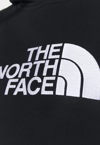 Sudadera negra con "THE NORTH FACE" en letras grandes y en negrita de color blanco, acompañada de un distintivo logo de medio domo blanco. Textura suave, diseño casual.
