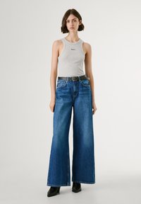 Graues ärmelloses Top mit kleinem Logo, kombiniert mit weiten blauen Jeans und einem schwarzen Gürtel. Schwarze High-Heel-Stiefel vervollständigen den Look.