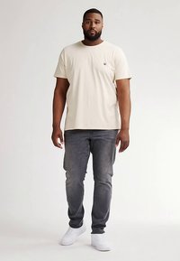 Tricou bej din bumbac, cu guler rotund și un mic logo pe pieptul stâng, asortat cu blugi gri din denim și adidași albi.