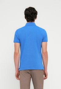 Μπλε κοντομάνικο πόλο μπλουζάκι με γιακά, με μονόχρωμη υφή. Φορεμένο με καφέ παντελόνι, παρουσιάζοντας ένα μινιμαλιστικό σχέδιο και εφαρμογή.