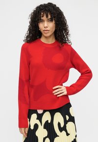 Κόκκινο πλεκτό πουλόβερ με στρογγυλή λαιμόκοψη, μακριά μανίκια και υφασματικές παραλλαγές υφής. Αντίθετα λουλουδάτα σχέδια σε πιο σκούρες κόκκινες αποχρώσεις.