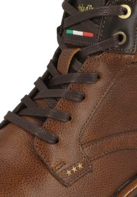 Stivale in pelle marrone con finitura texture, dotato di lacci marroni scuri, accentuato da stelle dorate e un emblema della bandiera italiana vicino al passante.