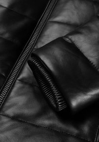 Close-up of black leather jacket sleeve with ribbed cuff and partially visible zipper on quilted leather body.