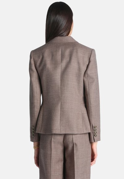 Femme aux longs cheveux foncés portant un blazer marron cintré et un pantalon assorti, vue de dos sur un fond uni.