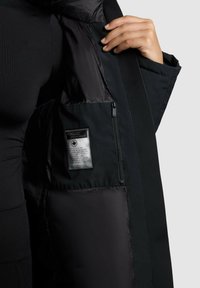 Zwarte parka met een soepel, glanzend interieur. Bevat een ritszak en een label met verzorgingsinstructies, geplaatst aan de linkerzijde.