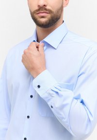 Camicia azzurra chiara realizzata in tessuto morbido, con colletto classico, chiusura anteriore con bottoni e taschino sul petto con bottoni neri.