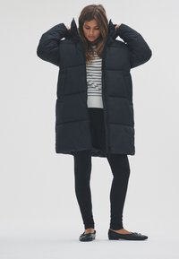 Fekete steppelt kabát kapucnival, elöl cipzárral és párnázott mintával. Csíkos felső és fekete leggingek fölött viselve.