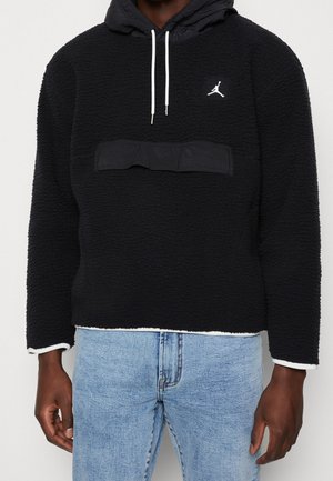 Mann trägt schwarzen strukturierten Hoodie mit weißen Kordeln und kleinem weißem Logo auf der Brust, kombiniert mit hellblauen Jeans.