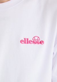 T-shirt en coton blanc avec un logo "ellesse" brodé en rose sur la partie gauche de la poitrine. Conception classique à col ras du cou avec une texture lisse.