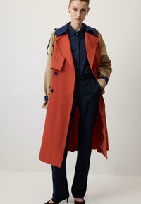 Abrigo tipo trench en tela naranja y beige con un diseño bicolor, gran cuello y acentos de botones, combinado con pantalones oscuros y una camisa azul marino.