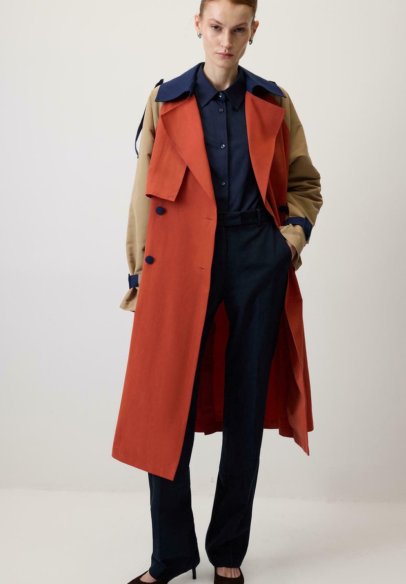 Abrigo tipo trench en tela naranja y beige con un diseño bicolor, gran cuello y acentos de botones, combinado con pantalones oscuros y una camisa azul marino.
