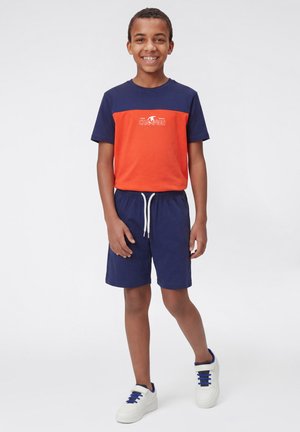 Lachende jongen staand, draagt een marineblauw en oranje Champion T-shirt, marineblauwe shorts met witte trekkoorden en witte sneakers met blauwe accenten.