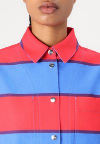 Punasilta punaisia ja sinisiä raitoja, kauluksellinen paita, jossa on hopeiset painonapit, kaksi etutaskua ja tekstuurinen kangas.