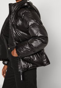 Veste doudoune noire avec une finition brillante, fermeture éclair et capuche. Présente des coutures diagonales et une étiquette de logo sur la tirette de la fermeture.