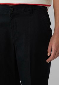Pantalons noirs ajustés en tissu léger avec une taille plate, dotés d'une fermeture à un bouton et poches latérales, présentés avec une main reposant à proximité.