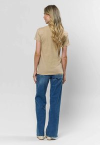 Béžové tričko s krátkymi rukávmi, v kombinácii s vysokými svetlomodrými džínsami, stojace na jednofarebnom pozadí.