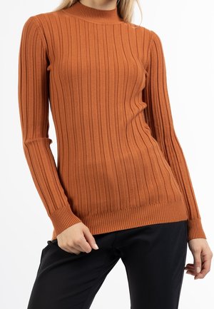 Femme portant un pull orange rouille côtelé manches longues à col montant et un pantalon noir sur fond blanc.