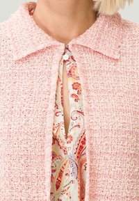 Giacca rosa con texture e colletto, dotata di chiusura a bottoni. Sotto, una blusa a fantasia in colori caldi è parzialmente visibile.