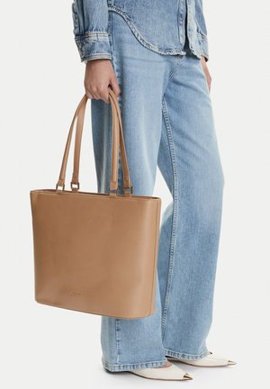 Persona sosteniendo un gran bolso tote de cuero marrón claro, con jeans azul claro, una chaqueta de mezclilla y zapatos blancos puntiagudos con puntas doradas.