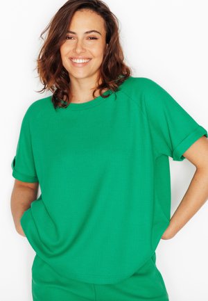 Donna sorridente con capelli castano ondulati, che indossa una maglia verde a maniche corte e pantaloni abbinati, in piedi con le mani nelle tasche.