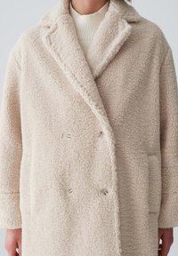 Beige doppelbreasted Mantel aus strukturiertem, flauschigem Material mit Reverskragen, zwei Fronttaschen und weißen Knopfakzenten.