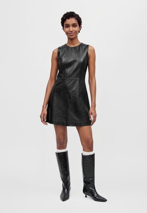 Modèle portant une mini-robe noire sans manches à effet cuir et des bottes noires montant jusqu’aux genoux, sur un fond clair uni.
