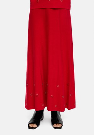Czerwona długa spódnica z srebrnymi ozdobami w postaci pierścieni blisko brzegu, zestawiona z czarnymi botkami na obcasie z odkrytymi palcami na białym tle.