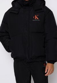 Svart pufferjacka med hög krage, framfickor och elastisk nederkant. Har en röd Calvin Klein Jeans-logotyp. Slät, blank yta.