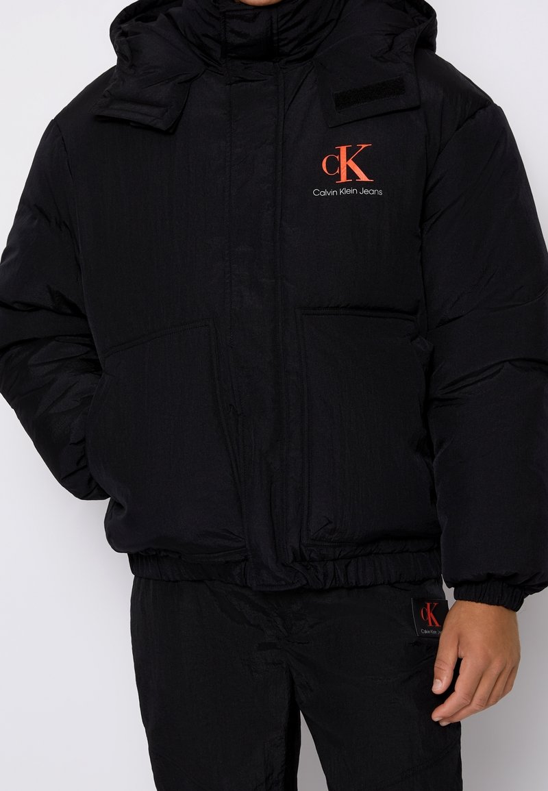 Svart pufferjacka med hög krage, framfickor och elastisk nederkant. Har en röd Calvin Klein Jeans-logotyp. Slät, blank yta.
