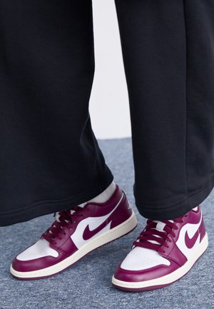 Personne portant un pantalon noir et des chaussettes blanches avec des baskets Nike bordeaux et blanches, debout sur un tapis gris.