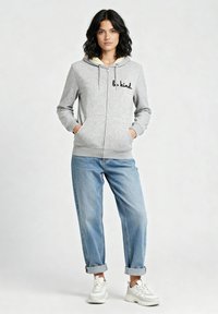 Felpa grigia con zip e testo "be kind" in nero, cappuccio foderato e due tasche frontali. Abbinata a jeans azzurri con risvolto e sneakers bianche.