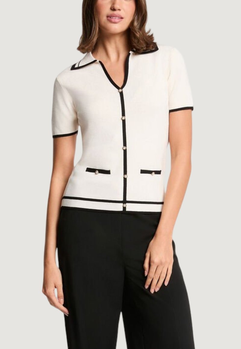 Woman wearing a white short-sleeve knit top with black trim, pearl buttons, and two decorative front pockets paired with black pants.