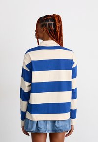 Personne avec des cheveux rouges tressés portant une chemise à rayures bleu et beige à manches longues et une jupe en denim courte, de dos sur un fond blanc.