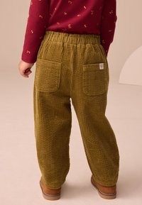 Olijfgroene corduroy broek met elastische tailleband, voorzien van twee achterzakken en een geribbelde structuur. Voet van een kind in een lichtbruine laars zichtbaar.