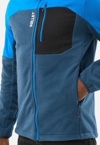 Fleece jacket in blue and black with a zippered chest pocket, contrasting zippers, and side pockets, featuring a soft texture and sporty design.