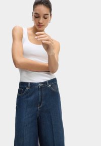 Dunkelblaue Jeans im weiten Bein mit hoher Taille, fünf Taschen-Design und goldfarbener Hardware. Kombiniert mit einem weißen Rippen-Top.