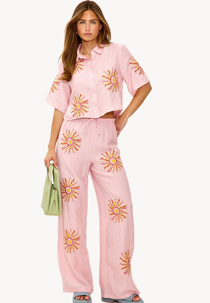 Lichtroze tweedelig outfit bestaande uit een cropped knoopshirt en wijde broek, beiden met zonbrodering in geel en oranje.