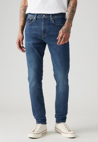 Blaue Denim-Jeans mit schlanker Passform, Fünf-Taschen-Design, Knopfverschluss und leichtem Wash-Effekt. Getragen mit weißen Turnschuhen.