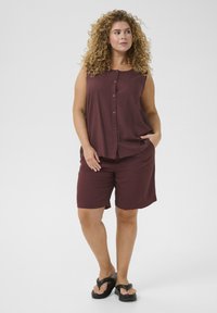 Top sin mangas de color marrón burdeos con botones frontales, combinado con pantalones cortos a juego. Tela suave, corte holgado y bolsillos laterales. Chanclas negras completan el look.