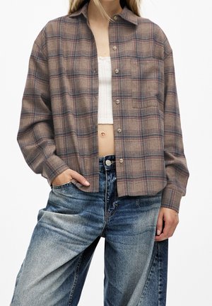 Femme portant une chemise à carreaux marron boutonnée sur un crop top blanc et un jean bleu délavé et ample, debout avec une main dans la poche.