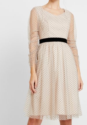 Beige knee-length dress with black polka dots, sheer long sleeves, sweetheart neckline, and black velvet waistband, worn by a standing person.