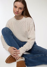 Sweter w kolorze kremowym o wzorze w warkocze, z prążkowanym dekoltem i teksturowanymi rękawami, w połączeniu z niebieskimi dżinsami i brązowymi butami na slip-on z sztucznym futerkiem.