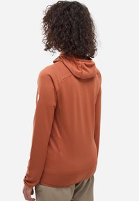 Haut à capuche long en couleur rouille, fabriqué en tissu texturé, avec une capuche et des poches latérales. Présente un motif subtil et une coupe ajustée.