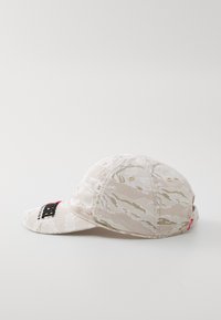 Cască bej cu un model de camuflaj ondulat, având un logo brodat pe față și un etichetă roșie pe lateral. Material moale și vizieră curbată.