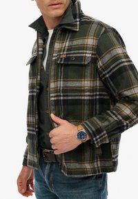 Giacca a quadri verde e nera con chiusura a zip, tasche sul petto e dettagli grigi; abbinata a un orologio blu e jeans di denim.