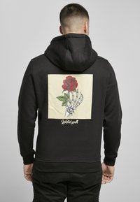 Felpa nera con cappuccio caratterizzata da una grafica sul retro: una rosa rossa tenuta da una mano scheletrica su uno sfondo di un quadrato crema. La scritta recita "Gioventù sprecata".